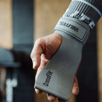 Hand wearing a velites gray wrist wrap with 'CrossFit' branding, blurred gym background