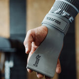 Hand wearing a velites gray wrist wrap with 'CrossFit' branding, blurred gym background