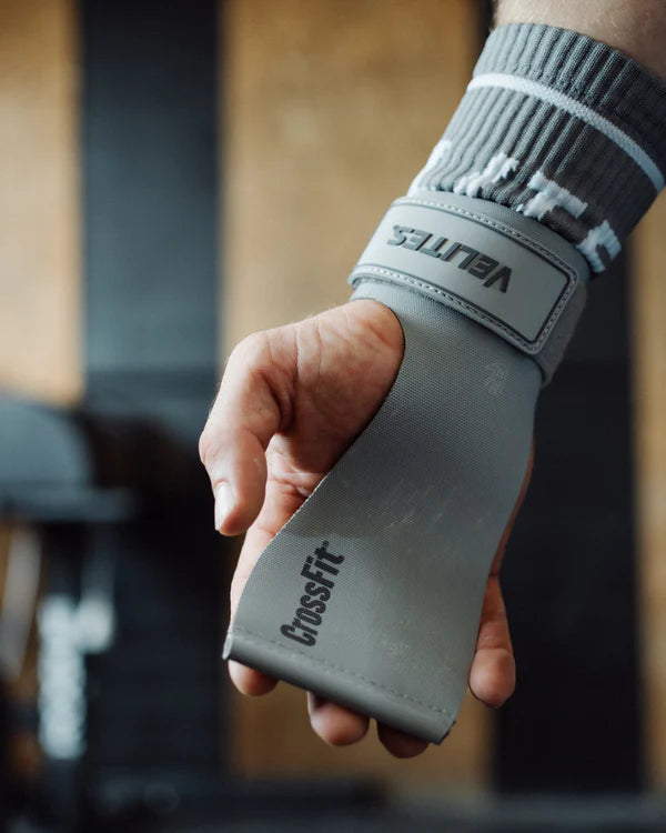 Hand wearing a velites gray wrist wrap with 'CrossFit' branding, blurred gym background