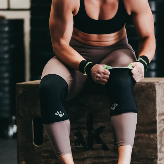 Woman sitting wearing frog knee sleeves on a wooden box in a gym setting
