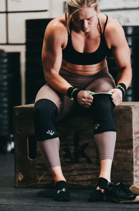 Woman sitting wearing frog knee sleeves on a wooden box in a gym setting